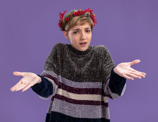 年轻不知所措的年轻漂亮女孩戴着圣诞花环看着镜头 在紫色的背景上孤立着空空的双手紫色显示圣诞快乐