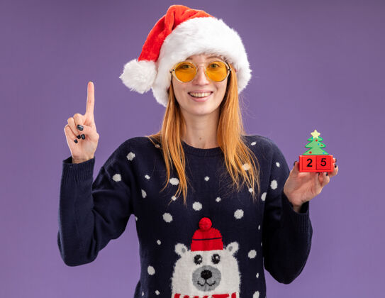 毛衣印象深刻的年轻漂亮的女孩穿着圣诞毛衣和帽子戴着眼镜拿着圣诞玩具点上孤立的紫色背景圣诞快乐女孩向上