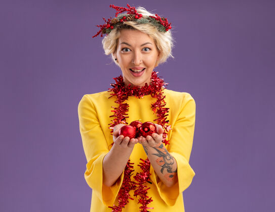 印象令人印象深刻的年轻金发女子头戴圣诞花环和金箔花环在脖子上看着相机举行圣诞饰品孤立在紫色的背景圣诞节花环紫色