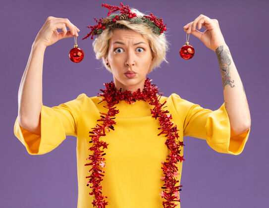 圣诞节不知所措的金发女郎头戴圣诞花环 脖子上戴着金属丝花环 看着相机拿着圣诞饰品 头上的紫色背景被孤立了女人不知所措饰品