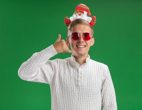 电话快乐的年轻帅哥戴着圣诞老人的头带 戴着眼镜 看着相机做着呼叫手势 隔离在绿色的背景上圣诞老人头带手势