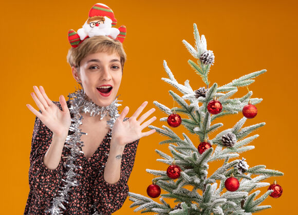 女孩年轻漂亮的女孩戴着圣诞老人的头带 脖子上戴着金箔花环 站在装饰好的圣诞树旁 两手空空的 孤立在橙色的背景上圣诞圣诞树显示