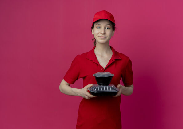 漂亮令人高兴的年轻漂亮的送货女孩穿着红色制服 戴着帽子 拿着食物容器 在深红色的背景上与复制空间隔离深红色女孩食物