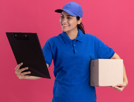 穿着面带微笑的年轻送货女孩穿着制服 手里拿着帽子盒 看着孤立在粉红色墙上的剪贴板盒子制服姿势