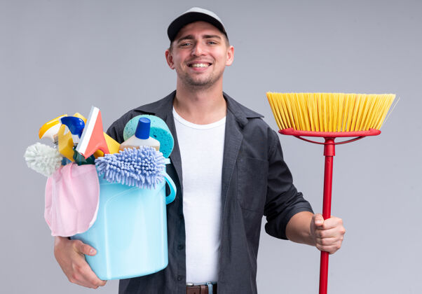 家伙面带微笑的年轻帅气的清洁工穿着t恤 戴着帽子 手里拿着一桶清洁工具 把拖把隔离在白色的墙上清洁桶工具