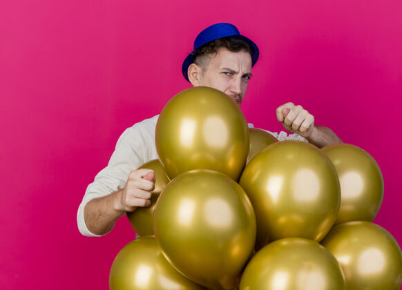 人皱眉的年轻英俊的斯拉夫党人戴着党的帽子站在气球后面看着前面做无花果标志隔离在粉红色的墙上皱眉人脸