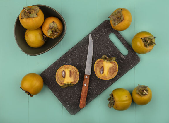 碗在蓝色的木桌上 一个碗上放着一把带有柿子水果的小刀 在黑色的菜板上放着橙色的圆形柿子水果的俯视图木板橘子柿子