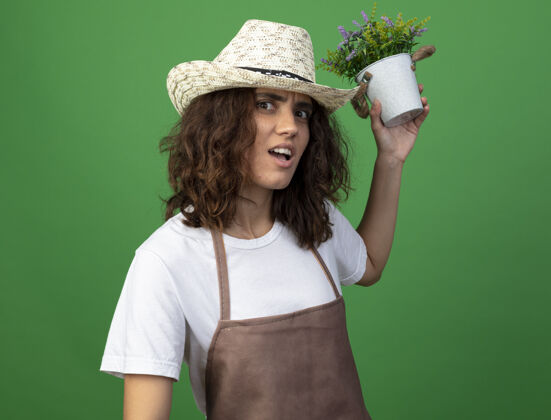 持有迷茫的年轻女园丁穿着制服 戴着园艺帽 在花盆里捧着花 背上隔离着绿色穿花盆花园
