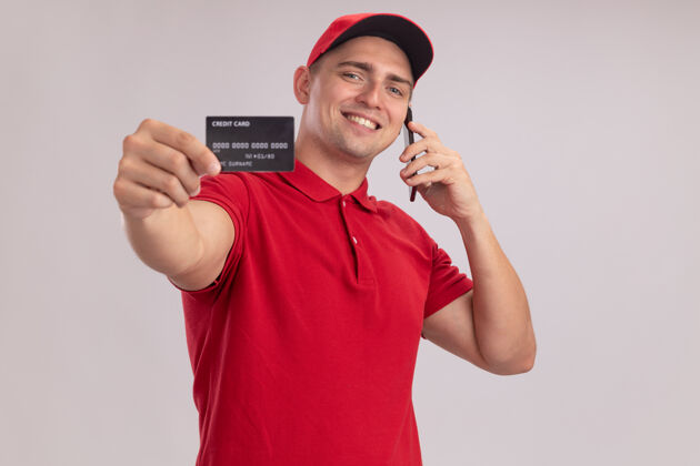 电话面带微笑的年轻送货员戴着制服 戴着帽子 举着信用卡 隔着白墙讲着电话送货持有男人