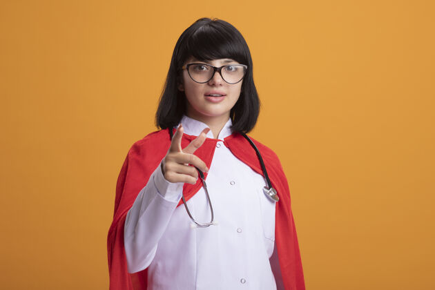 医疗年轻的超级英雄女孩戴着听诊器 穿着医用长袍和斗篷 戴着眼镜 在橙色的墙上显示着你们被隔离的姿势超级英雄眼镜表演