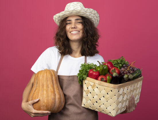 人年轻的女园丁面带微笑 穿着制服 戴着园艺帽 手里拿着一个蔬菜篮子 篮子里的南瓜是粉红色的粉红女人年轻