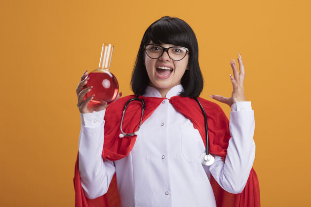 填补兴奋的年轻超级英雄女孩戴着听诊器 穿着医用长袍和斗篷 戴着眼镜 手里拿着装满红色液体的化学玻璃瓶蔓延液体人