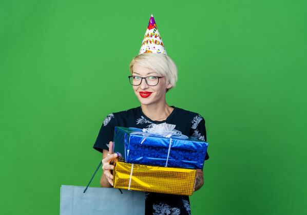 女孩年轻的金发派对女郎戴着眼镜 戴着生日帽 手里拿着纸袋和礼盒 看着前面隔离着的绿墙上有复印空间包人礼物