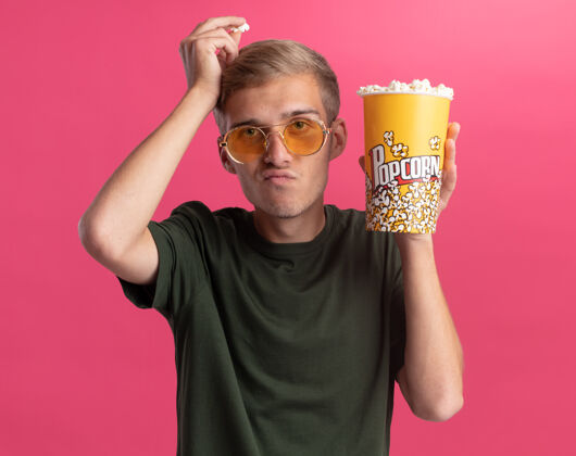 水桶穿着绿色衬衫戴着眼镜的年轻帅哥 手里拿着一桶爆米花 头上拿着一块爆米花 孤零零地站在粉红色的墙上抱着眼镜不愉快