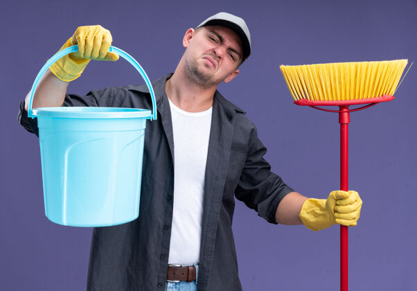 穿未出租的歪头年轻帅气的清洁工穿着t恤和帽子 戴着玻璃 拿着桶和拖把隔离在紫色的墙上帽子年轻拖把