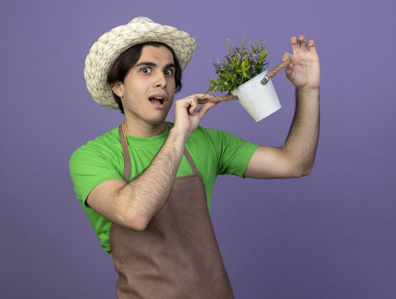 惊喜惊讶的年轻男园丁穿着制服戴着园艺帽捧着紫罗兰上孤立的花盆里的花花园花盆男性