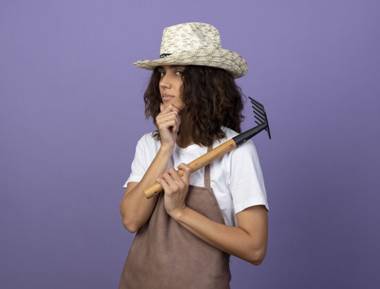 耙子自信的年轻女园丁穿着制服戴着园艺帽把耙子放在肩上 抓住下巴成人女人帽子