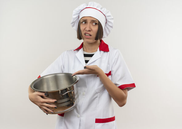 指着未出狱的年轻女厨师身着厨师制服 拿着锅 指着锅边看 在白色背景上隔离着复印空间不愉快年轻壁板