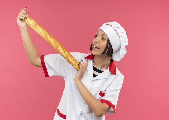烹饪快乐的年轻女厨师穿着厨师制服 拿着面包棒看着孤立的粉红色女欢乐年轻