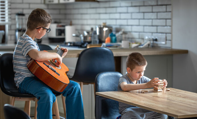 休闲一个小男孩弹吉他 他的哥哥在家的桌子旁用木块搭建了一个炮塔教育室内桌子
