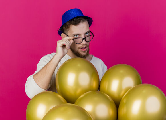 帅气年轻英俊的斯拉夫党人戴着党的帽子和眼镜站在气球后面抓着眼镜看着前面孤立的粉红色墙上人男人聚会