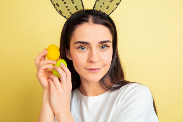 人类寻找彩蛋来了白人妇女作为一个复活节兔子在黄色的工作室背景复活节快乐的问候美丽的女模特人类情感的概念 面部表情 节日复制空间兔子明亮兔子