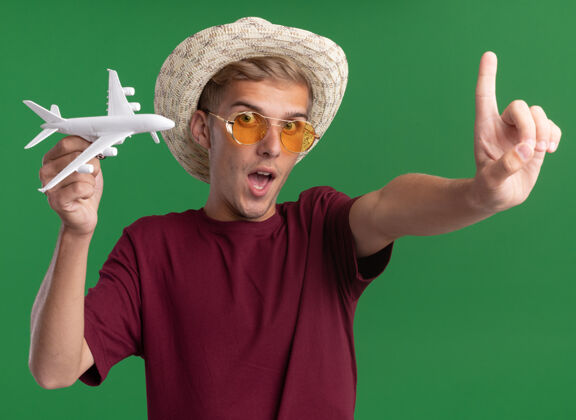 小伙子高兴的年轻帅哥穿着红衬衫戴着眼镜和帽子拿着玩具飞机伸出手指对着隔离在绿色墙上的相机衬衫拜托拿着