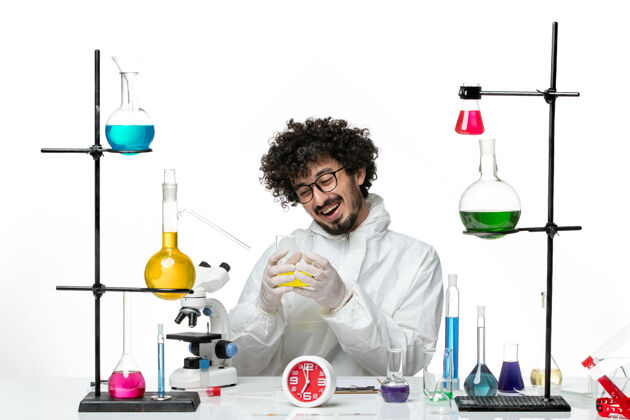 浅白色正面图穿着特殊套装的年轻男性科学家坐在浅白的墙上 拿着解决方案容器瓶子容器