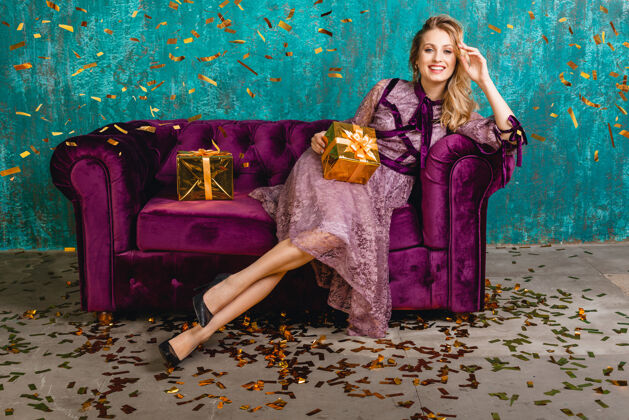 卷发穿着时尚紫罗兰色晚礼服的迷人女人坐在天鹅绒沙发上 带着礼物年轻乐趣女性
