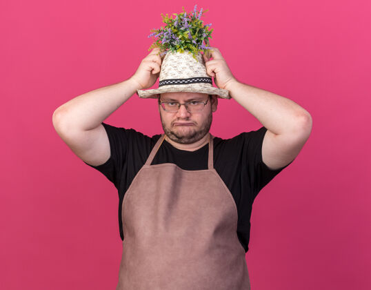 穿悲伤的年轻男园丁戴着园艺帽捧着花盆里的花 头孤立在粉红色的墙上悲伤年轻持有