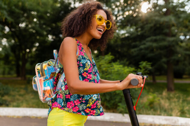 兴奋年轻时尚的黑人女子在公园里玩得很开心骑着电动脚踏车在夏天的时尚风格 五颜六色的时髦服装 穿着背包和黄色太阳镜黑色欢呼摩托车