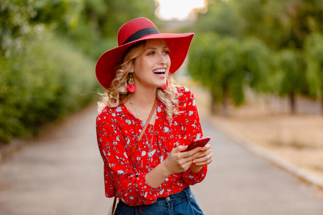 享受迷人的时尚金发微笑的女人在稻草红帽子和衬衫夏季时尚套装使用手机外表华丽装束