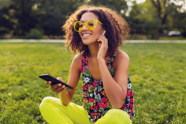 眼镜年轻时尚微笑的黑人女子用智能手机听音乐无线耳机在公园里玩得开心 夏日时尚多彩的风格 坐在草地上 戴着黄色太阳镜电话耳机兴奋