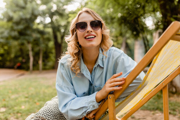 快乐漂亮的金发微笑的女人穿着时髦的衣服坐在躺椅上夏天时尚手提包