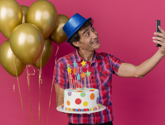 蛋糕微笑的中年白种人聚会男子戴着聚会帽站在气球旁拿着生日蛋糕在深红色背景下自拍帽子站着派对