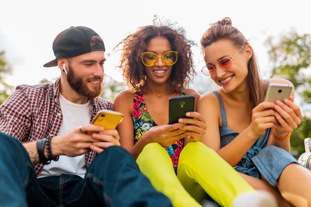 微笑快乐的年轻朋友们坐在公园里用智能手机微笑 男人和女人一起玩多彩音乐积极