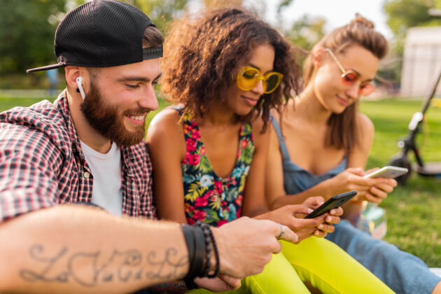 友谊快乐的年轻朋友们坐在公园里用智能手机微笑 男人和女人一起玩团体享受兴奋