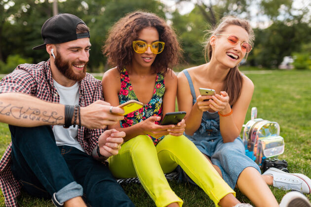 年轻快乐的年轻朋友们坐在公园里用智能手机微笑 男人和女人一起玩耳机朋友友谊