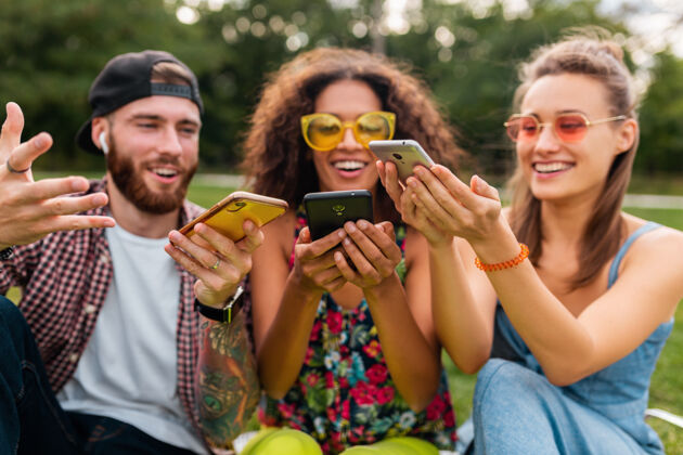 耳机快乐的年轻朋友们坐在公园里用智能手机微笑 男人和女人一起年轻多样性