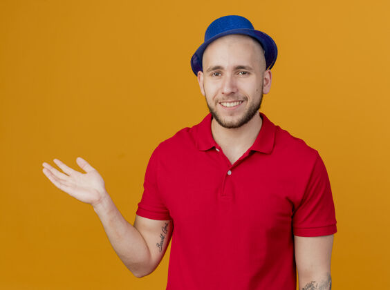 派对面带微笑的年轻英俊的斯拉夫党人戴着党的帽子看着相机显示空手孤立在橙色的背景帽子橙色表演