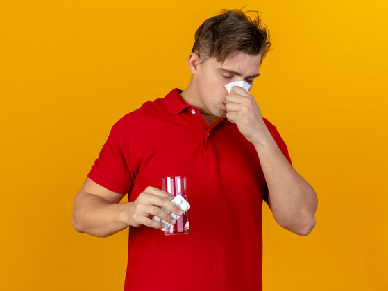 年轻人年轻英俊的金发病患拿着一包药片和一杯用餐巾擦鼻子的水 闭着眼睛隔离在橙色背景上橙色湿巾持有
