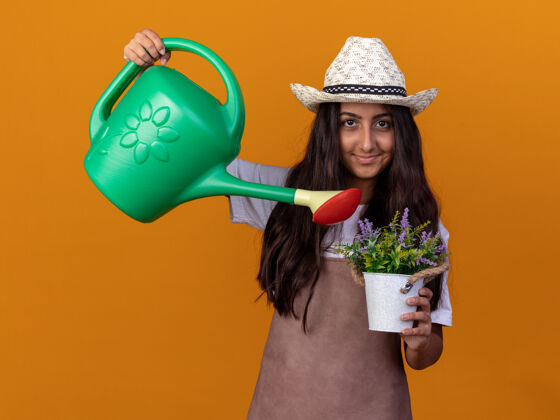 年轻快乐的年轻园丁女孩 围裙戴着夏帽 手里拿着浇灌罐 脸上带着微笑的盆栽浇灌植物站在橙色的墙上花园微笑站