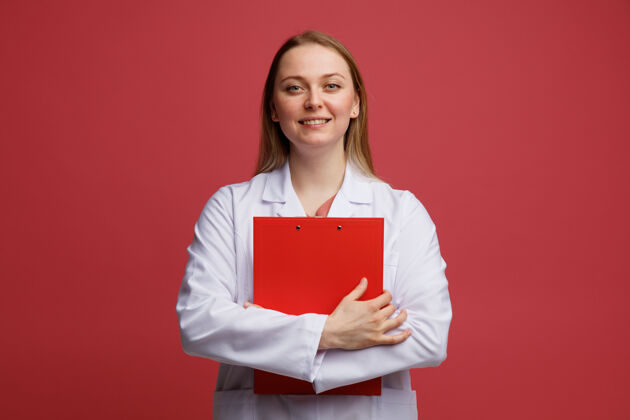 拥抱微笑的年轻金发女医生穿着医用长袍和听诊器围着脖子拥抱剪贴板周围医疗穿