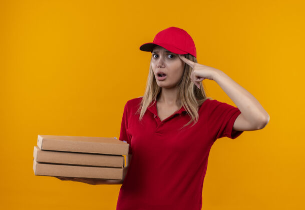 戴着困惑的年轻女送货员穿着红色制服 戴着帽子拿着披萨盒 手指放在额头上送货红色盒子