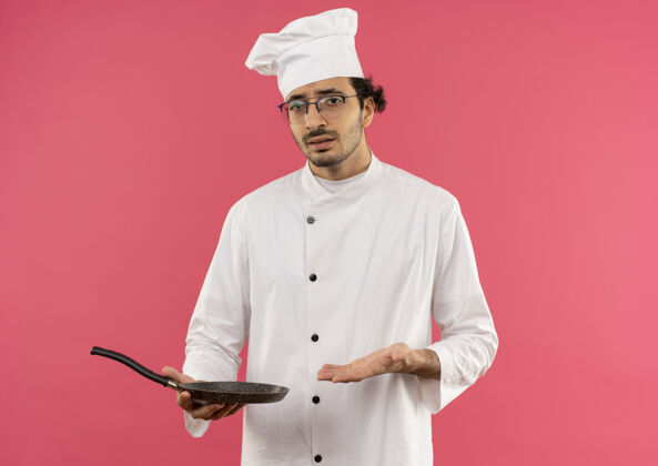 年轻困惑的年轻男厨师穿着厨师制服 戴着眼镜 手拿煎锅指着眼镜薯条平底锅