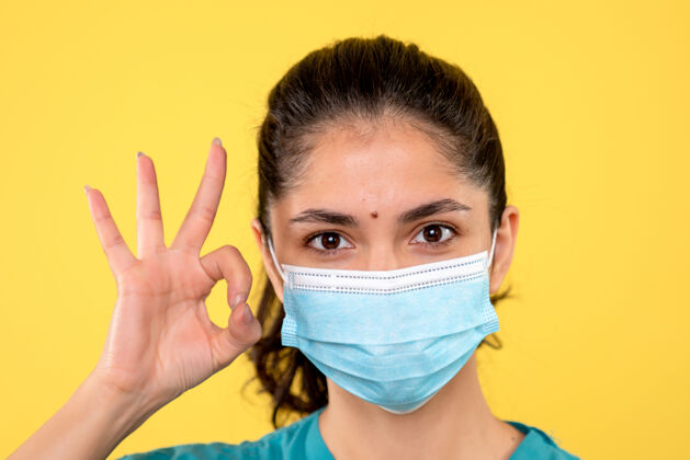 年轻女性戴着医用面罩的年轻女性的正面图 黄色墙上有okey标志面具漂亮保护封面