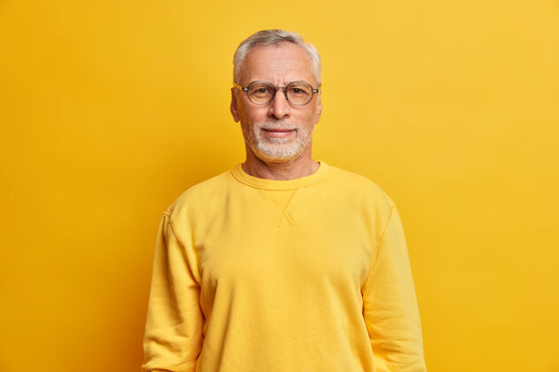时尚横拍的白发男人带着皱纹戴着眼镜 休闲的黄色套头衫直视前方 在室内摆出满意的表情姿势男性老年人皱眉