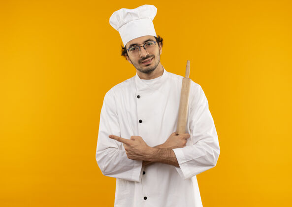 壁板高兴的年轻男厨师穿着厨师制服 戴着眼镜 拿着擀面杖 交叉着双手 指着隔离在黄色墙壁上的一侧 留着复印空间请男眼镜