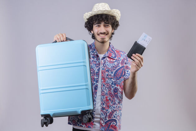 钱包微笑着的年轻英俊的卷发旅行者戴着帽子拿着钱包和飞机票和手提箱在与世隔绝的白墙上有复印空间年轻人机票穿
