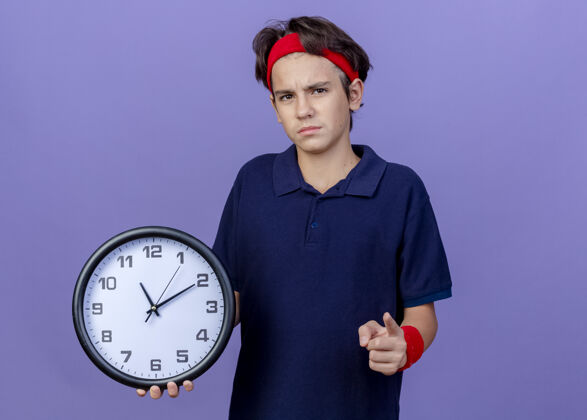 头带皱眉的年轻帅气的运动男孩戴着头带和护腕 戴着牙套 拿着时钟指针 隔离在紫色的墙上 留着复制空间腕带背带穿